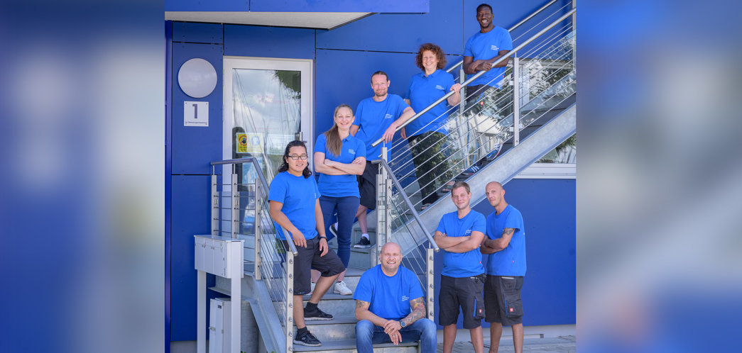 Gruppe von Männern und Frauen in blauen Shirts sind auf einer Treppe vor einer blauen Wand aufgestellt bei weichgezeichnetem Hintergrund